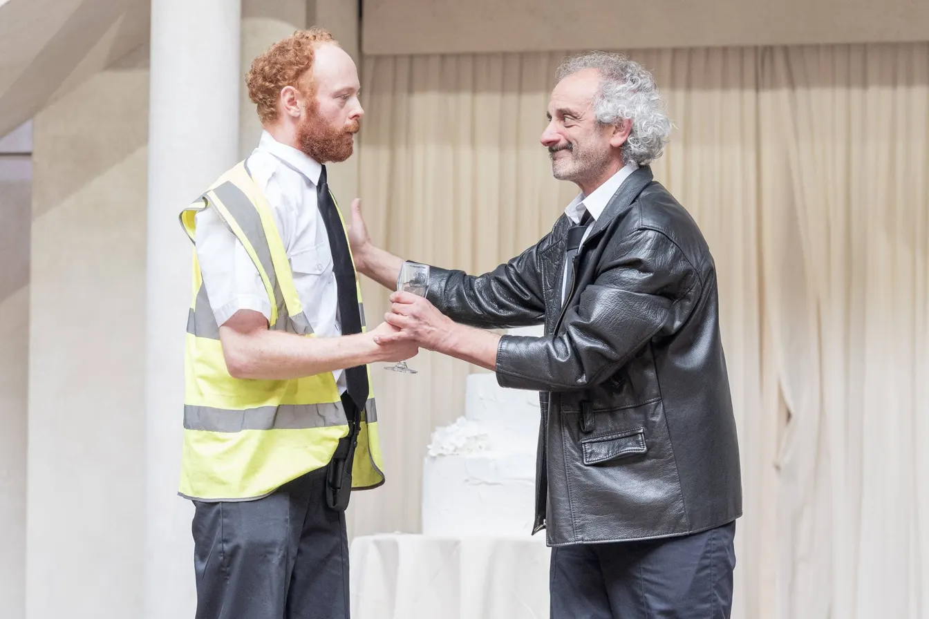 Two actors facing each other onstage, one wearing a high-visibility vest and the other a leather jacket, holding a glass.