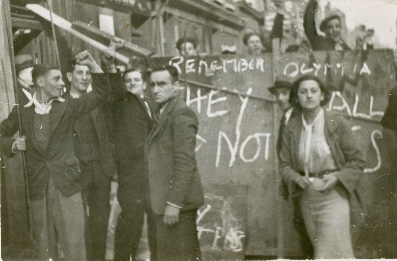 Black-and-white archive photograph of a crowd beside a handwritten protest banner, several raising their fists.