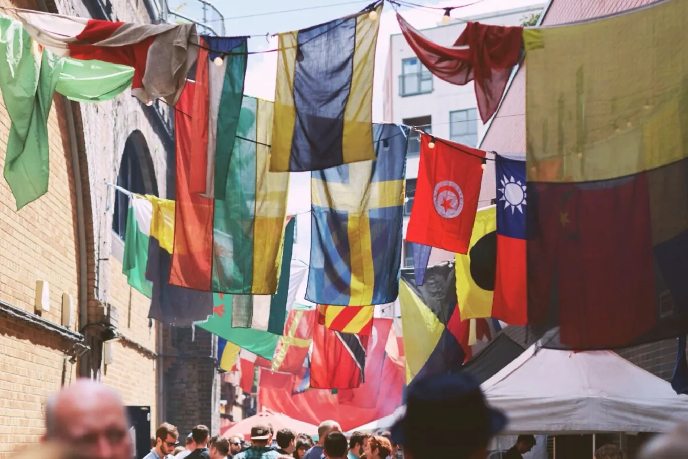 Colourful international flags hang above shoppers and market canopies in a narrow brick-lined lane.