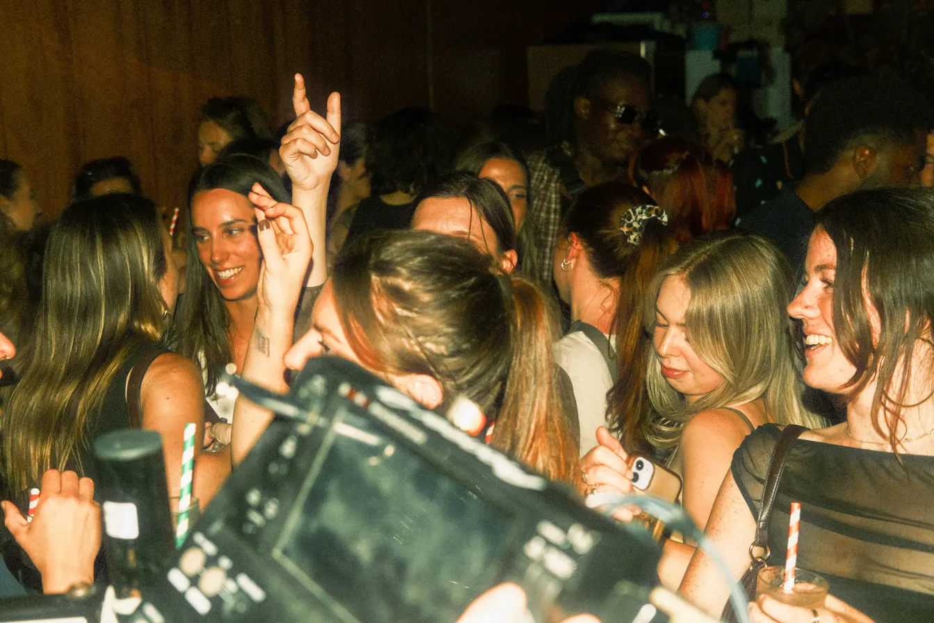 A crowded dance floor with smiling clubgoers holding drinks and raising their hands behind DJ equipment.